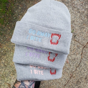 May include: Three gray knit beanies stacked, each with the embroidered phrase "GO AWAY I BITE" and a red fanged mouth design. The text and design colors vary: light blue, purple, and pink.
