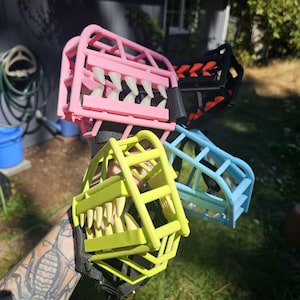 May include: A collection of four basket-style dog muzzles in pink, black, light blue, and neon yellow. Each muzzle features a rigid cage design with integrated, protruding plastic teeth along the front, secured by black nylon webbing straps.