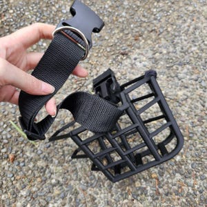 Può includere: Una museruola per cani a cestello di colore nero, caratterizzata da una gabbia rigida con motivo a griglia, cinghie in nylon intrecciato regolabili e una fibbia in plastica a sgancio laterale con anello a D in metallo.
