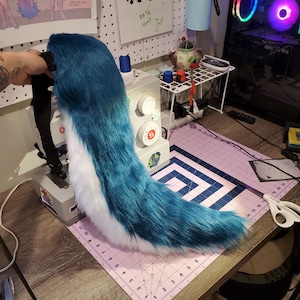 May include: A teal and white faux fur tail with a black strap. The tail is lying on a pink cutting mat with white grid lines.