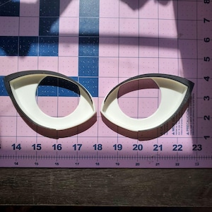 May include: Two eye-shaped craft stencils. Each stencil has a white interior and a black border. The stencils are placed on a pink and purple cutting mat with measurement markings in centimetres.