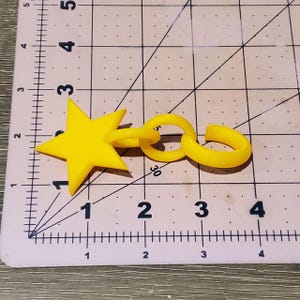 May include: A yellow plastic star-shaped charm attached to a chain with a loop at the end. The charm is about 2 inches wide and the chain is about 3 inches long.