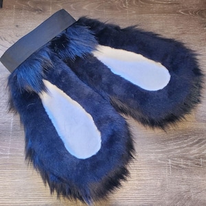 May include: A pair of black faux fur ears attached to a wide black elastic headband. The ears feature long, dark black outer fur with contrasting smooth, light-colored white inner panels, creating a distinct rounded shape for the accessory.