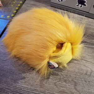Puede incluir: Un juguete de peluche amarillo con pelo largo y desgreñado. El juguete tiene una pequeña etiqueta adjunta.
