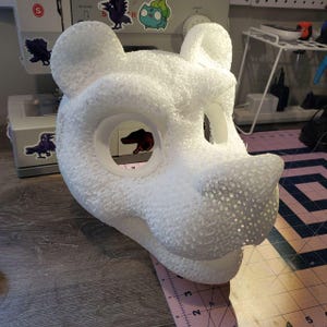 Könnte beinhalten: Weiße Schaumstoffmaske eines Tierkopfes mit Löchern für Augen und Nase. Die Maske ist texturiert und hat eine detaillierte Schnauze.