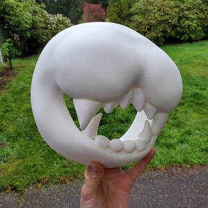 Può includere: Una scultura bianca e stilizzata di un teschio di creatura, con denti affilati e una forma arrotondata e aperta. La scultura è tenuta in mano su uno sfondo di erba verde e fogliame.