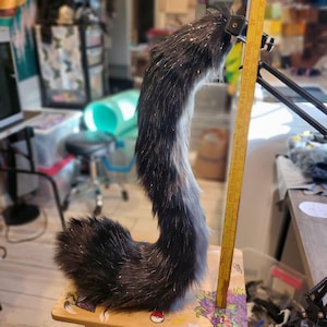 May include: A faux fur tail in shades of black and grey, with silver glitter accents. The tail is curved and appears to be approximately 61 cm long. It is resting on a wooden surface, with a measuring stick next to it.