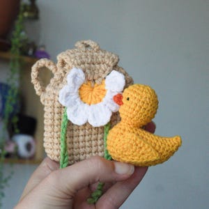 May include: A beige crocheted backpack featuring a large white and yellow daisy flower applique on the front, paired with a small, bright yellow crocheted duck toy. The backpack has a top handle and side straps, with green stems extending downward.