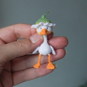 May include: A miniature crocheted duck figurine featuring a white body, orange beak and feet, and a green and white hat. The small handmade toy has embroidered black eyes and a detailed stitch pattern, showcasing a compact design.