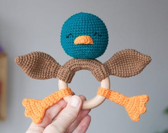 May include: A handmade crochet duck baby rattle. The duck has a teal head, brown wings, and orange beak and feet. The rattle is attached to a wooden ring. The duck's face has embroidered eyes and a smile.