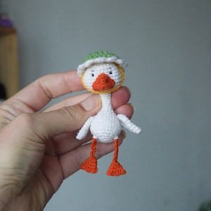 May include: A small, white crocheted duck toy featuring a bright orange beak and webbed feet. The duck wears a green hat with a white scalloped brim and has two small black eyes stitched onto its round, white head.