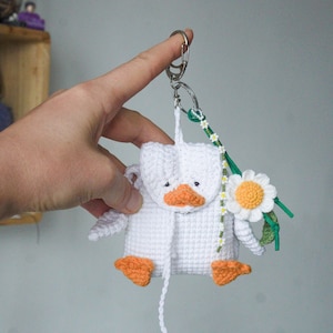 May include: A white crocheted duck-shaped pouch with an orange beak and feet, featuring a small flap closure. A metal keychain ring is attached to the top, adorned with a green cord, a string of tiny white daisy beads, and a larger crocheted daisy.
