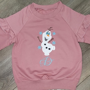 May include: A pink sweatshirt with ruffled sleeves and a crew neck. The front features an embroidered image of Olaf the snowman from Frozen, with blue snowflakes and the letter "B" in light blue script.