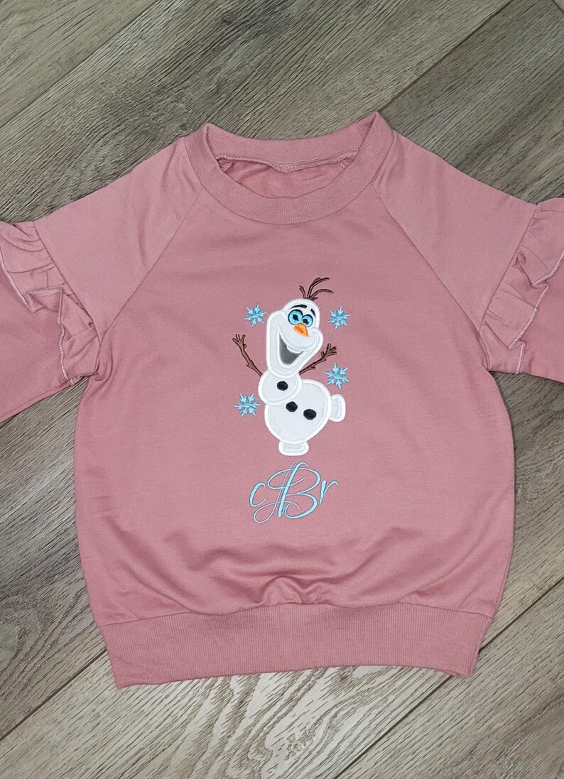 May include: A pink sweatshirt with ruffled sleeves and a crew neck. The front features an embroidered image of Olaf the snowman from Frozen, with blue snowflakes and the letter "B" in light blue script.