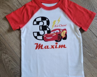 Op de afbeelding: Wit T-shirt met rode mouwen met een rode raceauto met nummer 3, een bliksem en de tekst "Ka-Chow!" De naam "Maxim" is onder de auto geborduurd.