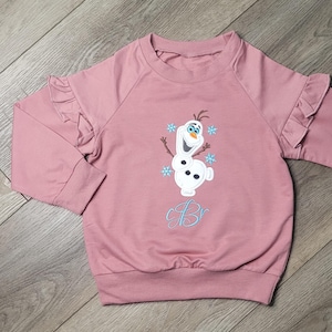 May include: A mauve-colored long-sleeved sweatshirt with ruffled sleeves. The front features an embroidered Olaf design with blue snowflakes and the initials "Br" in a light blue script. The sweatshirt has a ribbed hem and cuffs.