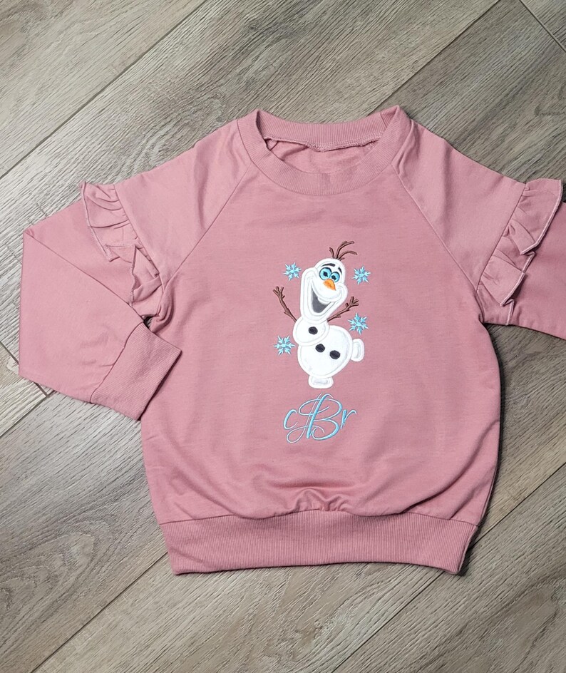 May include: A mauve-colored long-sleeved sweatshirt with ruffled sleeves. The front features an embroidered Olaf design with blue snowflakes and the initials "Br" in a light blue script. The sweatshirt has a ribbed hem and cuffs.