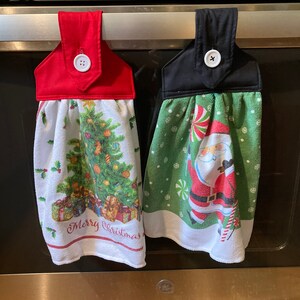May include: Two Christmas themed kitchen towels hanging on a silver oven rack. The towel on the left has a green Christmas tree with presents and the text "Merry Christmas". The towel on the right has a green background with a Santa Claus and candy canes. Both towels have a red or black fabric loop with a white button.