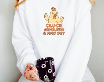 May include: White sweatshirt featuring a cartoon chicken with the text "CLUCK AROUND & FIND OUT" in orange. The chicken has a stern expression. A black mug with white flower designs is also visible.