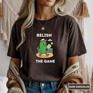 Può includere: T-shirt color cioccolato fondente con un cartone animato di cetriolo che gioca a pickleball. La maglietta ha le parole "RELISH THE GAME" stampate sopra e sotto la grafica del cetriolo. La maglietta è di vestibilità ampia.
