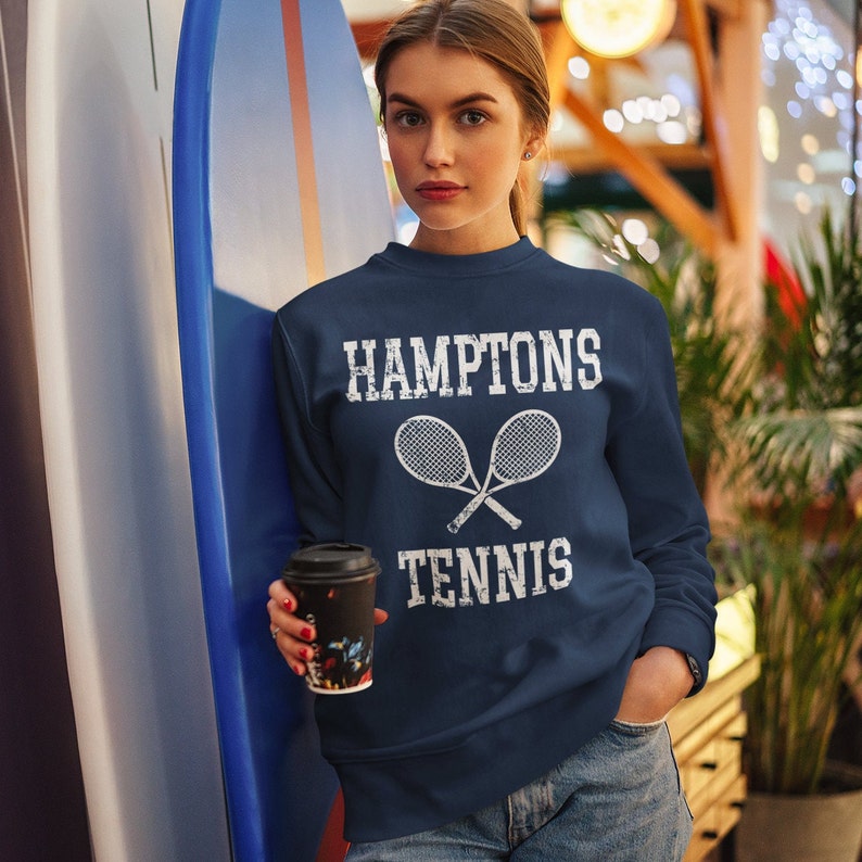 May include: A navy blue long-sleeve sweatshirt featuring white distressed text that reads HAMPTONS TENNIS, positioned above a graphic illustration of two crossed tennis rackets. The garment has a crew neckline and a standard fit.