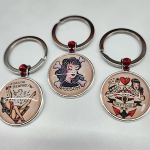 May include: Three silver keyrings with red rhinestone accents. Each keyring features a round, colourful, vintage-style tattoo design. The designs include a woman with the word "Poison" below her, a woman with the words "Sailor Beware" above her, and two women with the words "Screwed and Tattooed" below them.