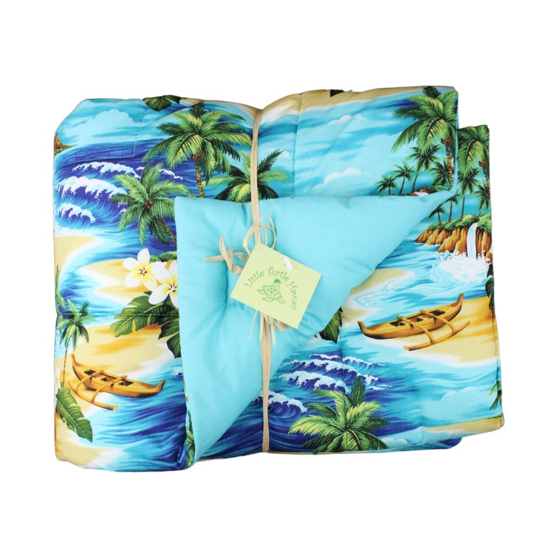 Puede incluir: Una colcha de estampado tropical azul y verde con un reverso turquesa. La colcha est&aacute; doblada y atada con una cuerda. Una peque&ntilde;a etiqueta con el texto "Little Turtle Haven" est&aacute; unida a la cuerda.