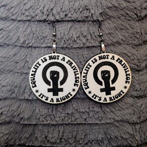 May include: A pair of wooden earrings with a white background and black text. The text on the earrings reads "Equality is not a privilege, it's a right." The earrings also feature a black fist inside a circle with a cross at the bottom.