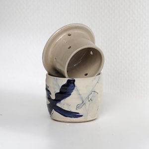 Pu&ograve; includere: Un set di burriera in ceramica a due pezzi con smalto color crema. La base presenta un motivo astratto di uccello dipinto a mano in blu scuro, mentre l'inserto superiore a forma di campana include piccoli fori circolari per l'uso.