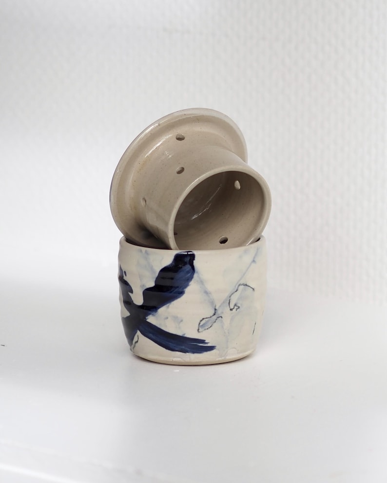 Pu&ograve; includere: Un set di burriera in ceramica a due pezzi con smalto color crema. La base presenta un motivo astratto di uccello dipinto a mano in blu scuro, mentre l'inserto superiore a forma di campana include piccoli fori circolari per l'uso.
