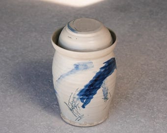 Può includere: Un barattolo cilindrico in ceramica con coperchio abbinato, caratterizzato da una base color crema con pennellate blu scuro dipinte a mano e dettagli a linee sottili. Il contenitore ha un corpo leggermente affusolato e una finitura smaltata.