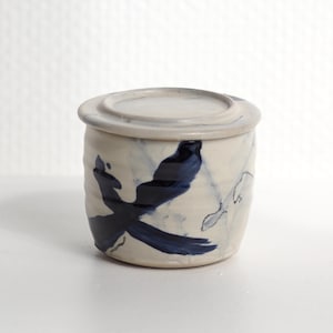 Pu&ograve; includere: Un barattolo cilindrico in ceramica con coperchio abbinato, caratterizzato da un colore di base bianco sporco e motivi a pennellate blu scuro dipinti a mano. La superficie esterna presenta una sottile trama a coste e una finitura smaltata.