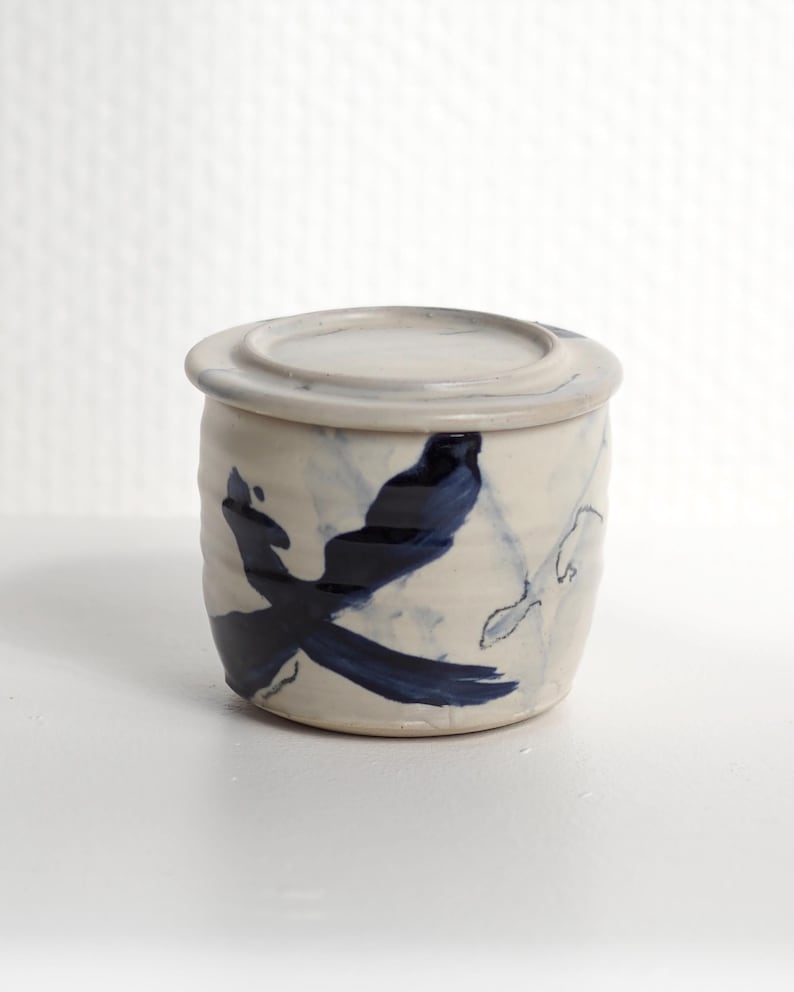 Pu&ograve; includere: Un barattolo cilindrico in ceramica con coperchio abbinato, caratterizzato da un colore di base bianco sporco e motivi a pennellate blu scuro dipinti a mano. La superficie esterna presenta una sottile trama a coste e una finitura smaltata.
