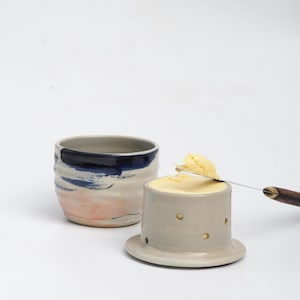 Pu&ograve; includere: Un set di burriera in ceramica composto da una base grigio chiaro con piccoli ritagli circolari e una ciotola abbinata con pennellate blu e color pesca. Un coltello da burro in metallo con manico in legno poggia sul burro.