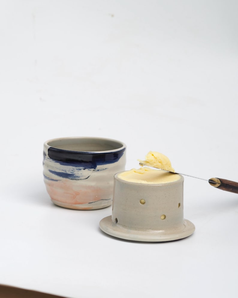 Pu&ograve; includere: Un set di burriera in ceramica composto da una base grigio chiaro con piccoli ritagli circolari e una ciotola abbinata con pennellate blu e color pesca. Un coltello da burro in metallo con manico in legno poggia sul burro.