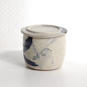 Pu&ograve; includere: Un piccolo barattolo cilindrico in ceramica con coperchio abbinato, caratterizzato da una base color bianco sporco e pennellate astratte blu scuro dipinte a mano. La superficie esterna presenta sottili scanalature orizzontali e una finitura smaltata.