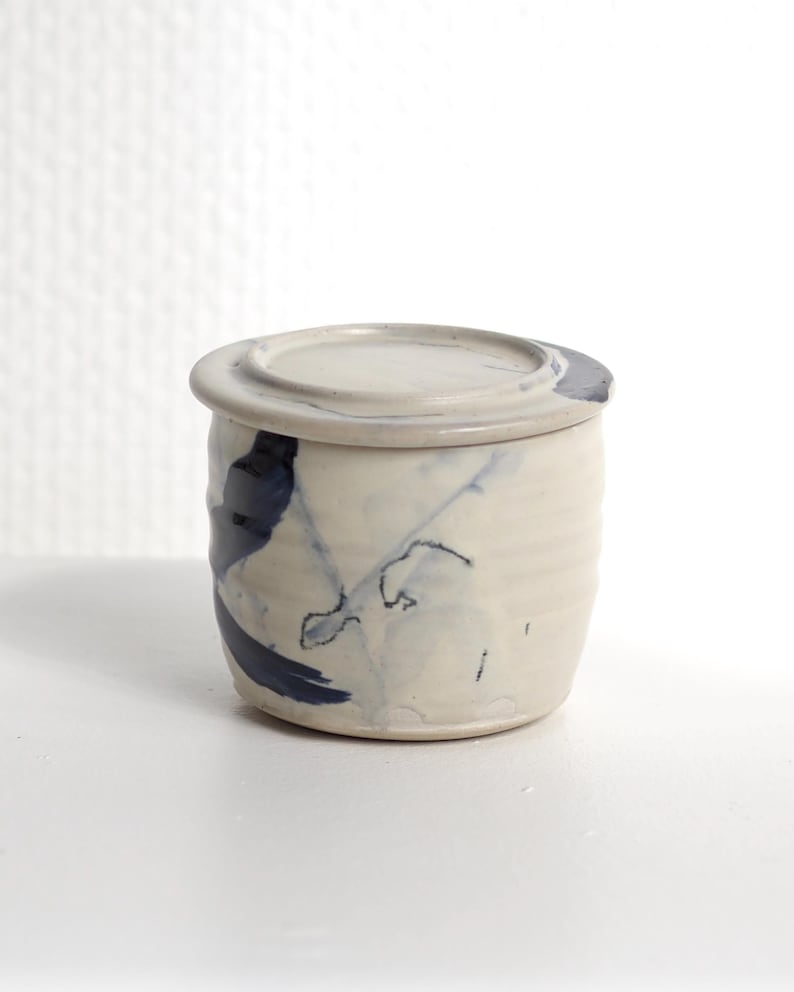Pu&ograve; includere: Un piccolo barattolo cilindrico in ceramica con coperchio abbinato, caratterizzato da una base color bianco sporco e pennellate astratte blu scuro dipinte a mano. La superficie esterna presenta sottili scanalature orizzontali e una finitura smaltata.
