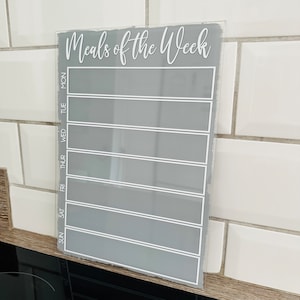 Op de afbeelding: Een grijs acryl droog uitwisbaar bord met een witte rand en de tekst "Meals of the Week" bovenaan. Het bord heeft zeven rijen voor elke dag van de week, gelabeld met de afkortingen "Sun", "Mon", "Tue", "Wed", "Thur", "Fri" en "Sat".