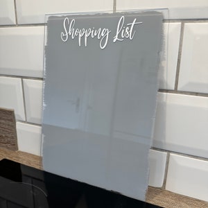May include: A clear acrylic shopping list board with the words "Shopping List" in a white script font. The board has a grey painted section and a textured edge. The background features white tiles.