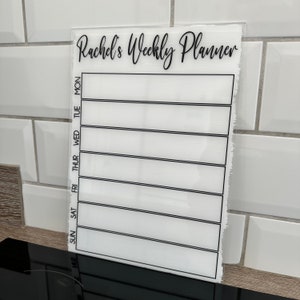 May include: White acrylic weekly planner with black text that reads "Rachel's Weekly Planner". The planner has seven columns for each day of the week, with the days of the week labelled in black text.