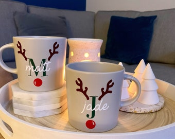 May include: Two gray ceramic mugs with reindeer antlers and red noses. The mugs are personalized with the names "Matt" and "Jade".