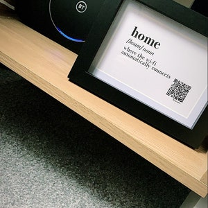 May include: A black framed picture with the word "home" defined as "[hoʊm] noun where the wi-fi automatically connects" and a QR code. The picture is sitting on a wooden shelf with a black speaker with a blue light ring.