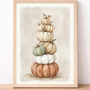 Puede incluir: Una impresión en acuarela enmarcada que presenta una pila vertical de siete calabazas de varios tamaños y colores, incluyendo tonos naranja, blanco, verde salvia y beige, sobre un fondo neutro de tonos suaves en un marco de madera clara.