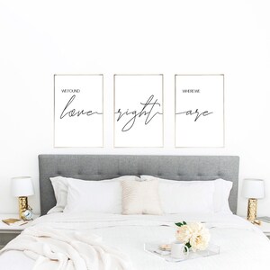 Può includere: Tre stampe incorniciate in bianco e nero con il testo "We found love right where we are" in un carattere corsivo. Le stampe sono appese a una parete bianca sopra una testiera grigia con un copriletto bianco.