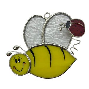 May include: A stained glass bee with a yellow body, black stripes, and a white face. The bee has a ladybug on its back with red wings and black spots.
