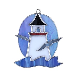 May include: A stained glass ornament depicting a white lighthouse with a red roof and black trim, set against a blue and purple sky with two white birds flying overhead. The lighthouse is surrounded by blue waves.