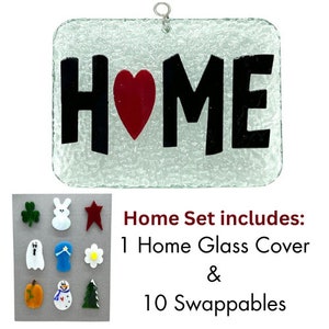 May include: A glass home sign with a red heart and the word "HOME" in black lettering. The sign comes with 10 interchangeable glass pieces, including a shamrock, bunny, star, ghost, hat, flower, pumpkin, snowman, and tree.