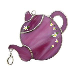 May include: A purple stained glass teapot with a silver trim and white daisy accents. The teapot has a small crystal drop hanging from the spout.