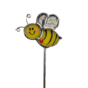 May include: A yellow and black bee with a smiling face and a red heart on its abdomen. The bee has clear wings and is attached to a silver metal stick.