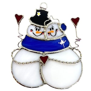 May include: A stained glass ornament depicting two snowmen holding hands and wearing a blue scarf. They are each holding a red heart.